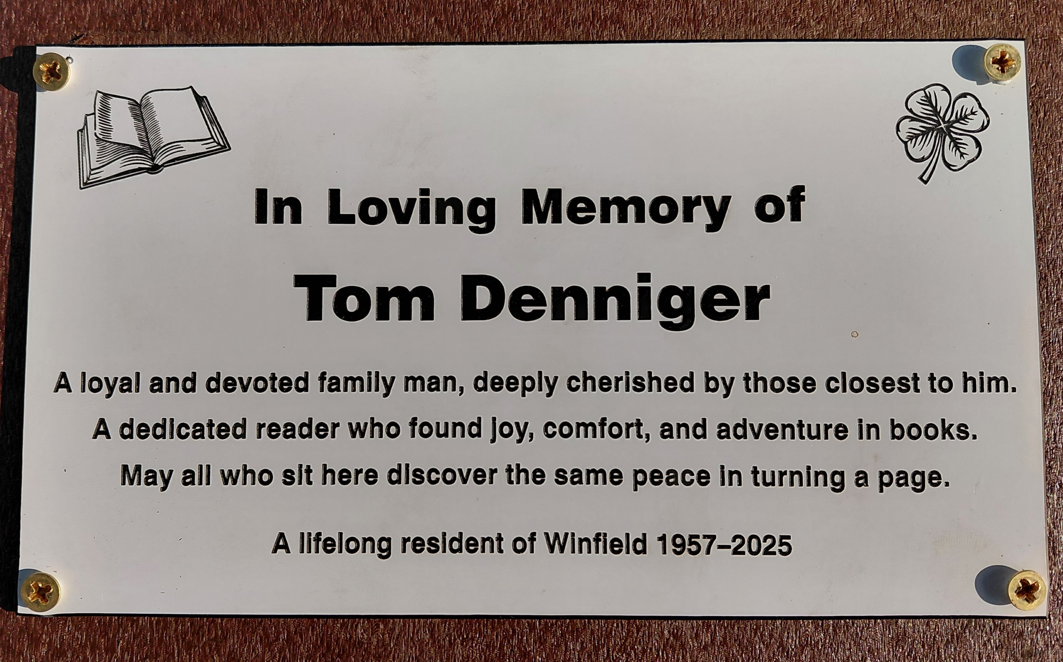 Plaque reading "In loving memory of Tom Denniger. A loyal and devoted family man, deeply cherished by those closest to him. A dedicated reader who found joy, comfort, and adventure in books. May all who sit here discover the same peace in turning a page. A lifelong resident of Winfield 1957-2025.